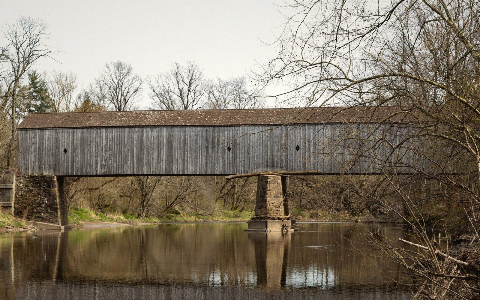A weathered wooden covered bridge spans a calm river, supported by stone piers